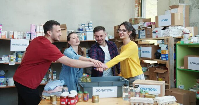Happy group of young volunteers in casual attire stacking their hands together after successful teamwork at charity storage. Concept of unity, support and humanitarian aid.