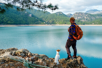 A man and a jack russell terrier enjoying the scenic view of mountain lake. Traveling with the dog concept. Hipster guy backpacking with his pet pup. Background, copy space.