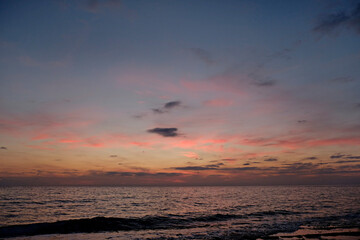 Fototapeta premium Beautiful seascape of sun setting over calm water with wind patterns and colorful pink spindrift clouds in the sky. Ocean view with horizon, background with a lot of copy space for text.