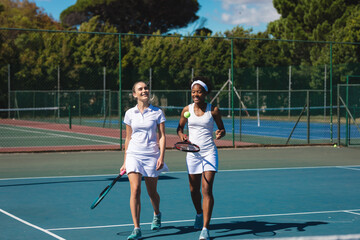 Full length of smiling biracial female doubles team walking at tennis court on sunny day