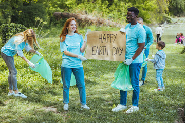 Diverse Group of People Picking Up Trash in The Park Volunteer Community Service. Happy international volunteers holding placard with 'happy Earth day' message.