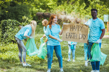 Diverse Group of People Picking Up Trash in The Park Volunteer Community Service. Happy international volunteers holding placard with 'happy Earth day' message.