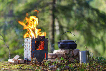A burning camping wood stove with a kettle, a mug and firewood on the background of the forest....