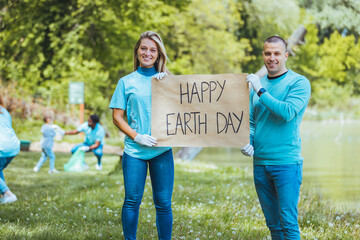 Diverse Group of People Picking Up Trash in The Park Volunteer Community Service. Happy international volunteers holding placard with 'happy Earth day' message.