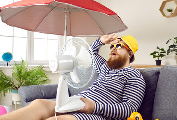 Funny sweaty chubby bearded man in swimsuit sitting at home, suffering from crazy summer heat, wiping sweat off forehead, holding electric fan, wishing for heatwave to stop and fresh breeze to blow - obrazy, fototapety, plakaty