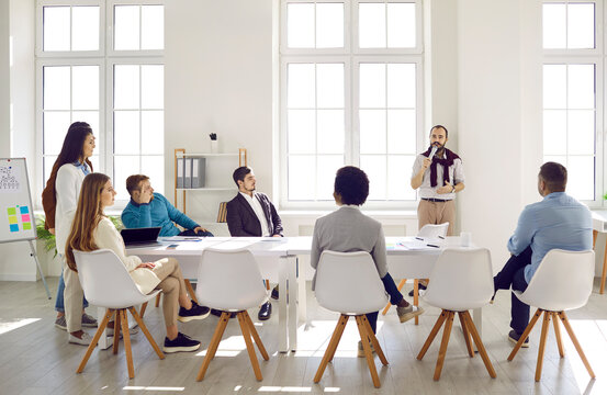 Team of people listening to experienced business coach during meeting in modern light office. Young man standing in front of diverse group of men and women, holding microphone and speaking