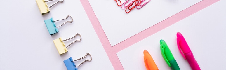 flat lay of colorful fold back clips, paper clips and pens on white papers on pink, banner.