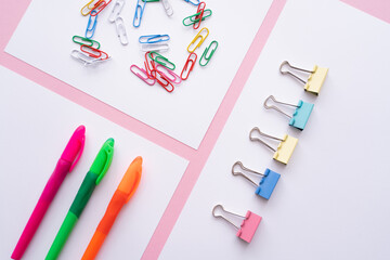 flat lay of colorful fold back clips, paper clips and pens on white papers on pink.