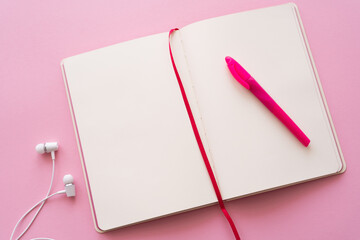 top view of open notebook and pen near wired earphones on pink.