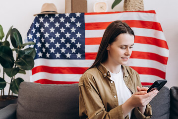 Beautiful smiling young woman sitting on cozy sofa at home with USA flag, cute happy caucasian female 20s old years with smartphone looking Independence Day celebration. Patriotic US holiday concept