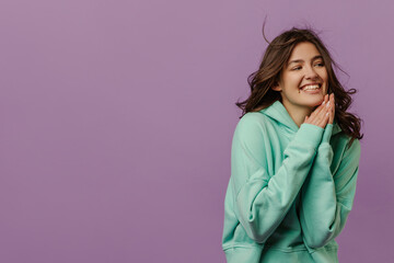 Happy young caucasian girl is smiling away in purple background with room to spare. Brunette girl wear turquoise sweatshirt holding her hands together. Mood, lifestyle, concept .