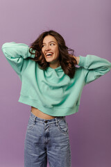 Happy caucasian young girl with wavy dark hair smiling looking away on purple background. Pretty woman wears turquoise hoodie and jeans.