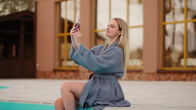 pretty woman in gown is resting in spa hotel with open swimming pool, taking selfie by smartphone