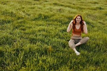 Young woman with red hair is happy and smiling sitting on the fresh grass in the park in the sun