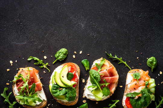 Open sandwiches set with cream cheese, prosciutto, salmon, avocado and fresh greens and vegetables. Top view at dark background with copy space.