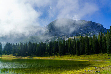 Ein idyllischer Bergsee liegt eingebettet in eine saftig grüne Wiese, während sich dichte Nadelwälder vor einem imposanten, teilweise in Wolken gehüllten Gebirgsmassiv unter blauem Himmel erstrecken.