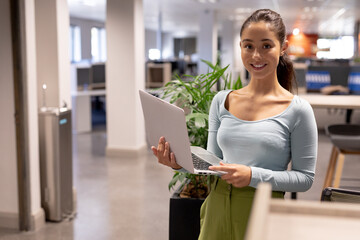Portrait of smiling young female hispanic advisor holding laptop while working at modern workplace