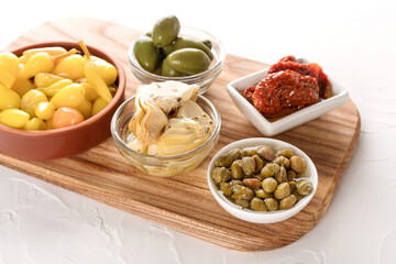 Wooden serving board with selection of antipasti on white stone table background - pickled artichokes, capers, baby peppers, sun dried tomatoes and olives. Selective focus