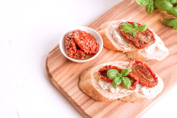 Top view on delicious toasts with ricotta cheese, sliced sun dried tomatoes topped with basil and dried tomatoes with olive oil in a bowl on wooden serving board. Classic italian antipasti. Copy space