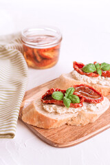 Vertical image of delicious toasts with ricotta cheese, sliced sun dried tomatoes topped with basil and dried tomatoes with olive oil in jar on wooden board, white table. Classic italian appetizer