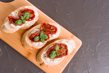 Bruschettas or toasts with soft ricotta cheese and sliced sun dried tomatoes topped with basil on wooden cutting board, concrete table background. Traditional Italian appetizer, top view