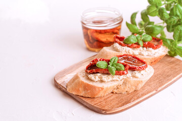 Delicious toasts with ricotta cheese, sliced sun dried tomatoes topped with basil and dried tomatoes with olive oil in jar on wooden serving board, white table background. Classic italian appetizer