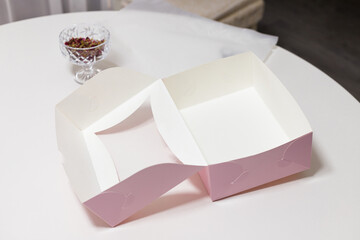 Pink empty paper box for sweet cakes, cupcakes or muffins on white kitchen table. Vase of dried rose flower petals on background