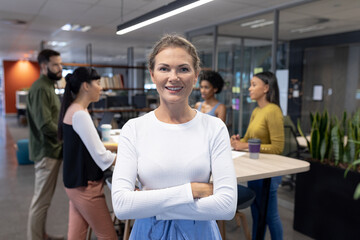 Portrait of smiling caucasian female advisor with arms crossed against team at workplace