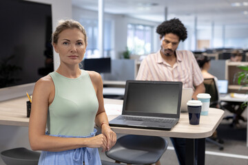 Portrait of confident cauasian businesswoman standing by laptop against biracial businesswoman
