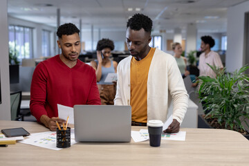 Biracial businessmen discussing over document with laptop at desk in modern workplace