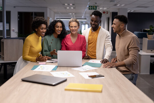 Smiling biracial businesswomen and businessmen discussing over laptop during meeting in workplace