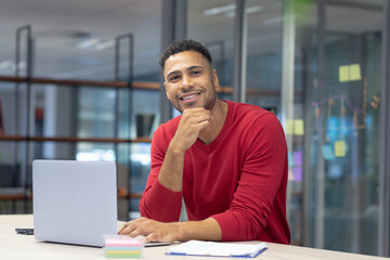 Portrait of happy hispanic male advisor sitting with laptop and document at modern workplace