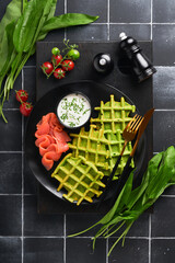 Green Belgian waffles. Spinach or wild garlic or pesto waffles with red salmon and cream sauce on old black tile table background. Delicious breakfast, snack, brunch on dark background, top view.
