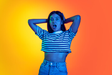 Portrait of young emotive girl with shocked facial expression, holding hands behind head isolated over orange yellow background in neon light