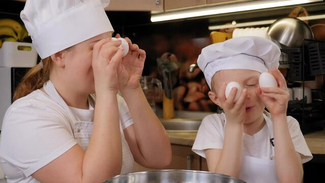 Close-up Portrait Of A Mother And Child In An Apron And Caps In The Kitchen, They Are Holding Eggs In Front Of Their Eyes, Smiling And Looking At The Camera.