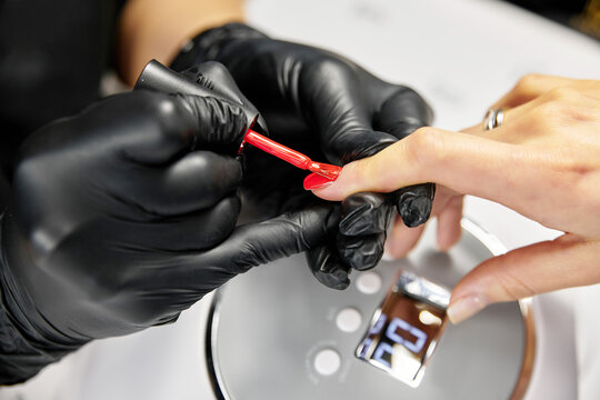 the master makes a manicure with a gel polish coating for a woman with clean nails. a master in black latex gloves paints a nail with red varnish