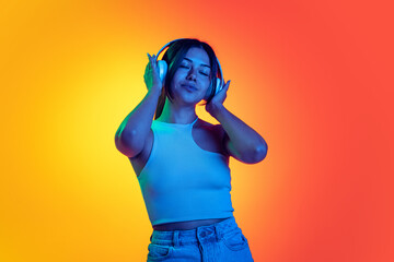 One young calm girl listening to music in heaphones with eyes closed isolated over orange yellow background in neon light