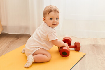 Full length indoor shot of charming cute infant baby crawling on yoga mat with dumbbells, exploring sport equipment, wearing bodysuit.