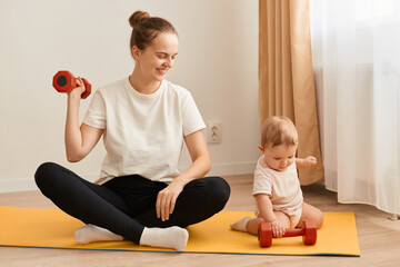 Indoor shot of smiling woman sitting on yoga mat with her baby daughter and training her arms, her...