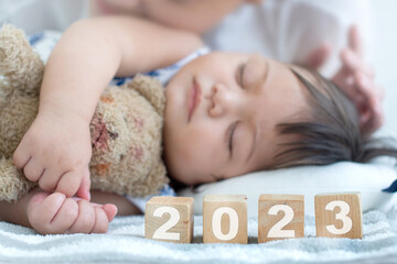 Close up, Happy attractive young mother with her 6 month old daughter sleeping, woman places the number 2023 wooden block, selective focus