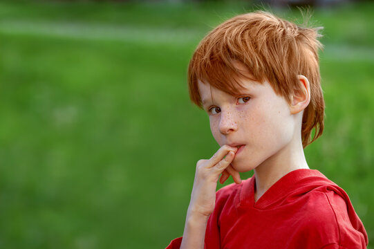 A brooding boy-child in a red T-shirt bites his nails outdoors. The concept of children in a difficult situation, parents' divorce, stress.