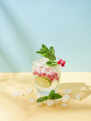 Non-alcoholic soda water tonic. Cold drink in crystal glass with ice, lime slice, redcurrant and mint lefs on yellow and blue background.