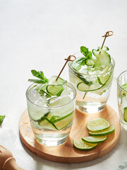 cold infused cucumber water in glasses with ice, lime and thin slices of cucumber skewered. Close-up view with gray background.