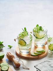 Glasses with fresh cucumber water garnished with thin, wavy cucumber strips on a skewer. Round wooden board and lime slices.