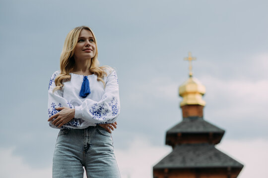 Young woman stands in Ukrainian embroidered shirt against background of church dome and sky.
