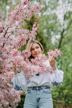 Young blonde woman walks in garden and enjoys by blooming sakura trees.