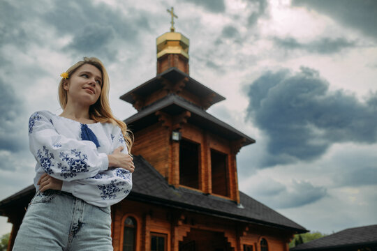 Young woman stands in Ukrainian embroidered shirt against background of church and sky.