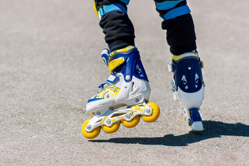 Fototapeta na wymiar A little child rides blue yellow and white roller skates. Cropped shot of a kid learning to rollerblade on asphalt. Sunny summer day