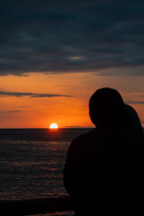 Rear view of the silhouette of a couple in love standing on the pier and enjoying the stunning sunset of the setting sun over the horizon of the sea. Vertical photo
