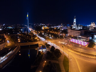 Flag of Ukraine on illuminated river embankment with water reflection at night. City aerial view above river Lopan near Dormition Cathedral in Kharkiv, Ukraine with long exposure